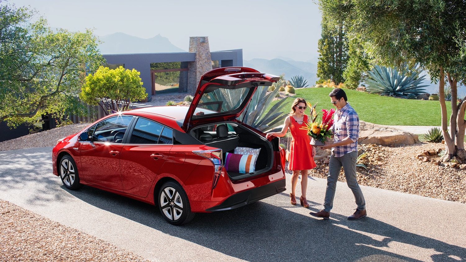 Image of a couple with sunglasses putting a pot of flowers into the trunk of a red Toyota Prius.