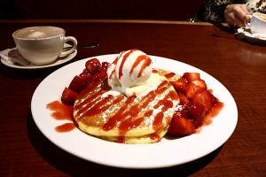 pancakes on a plate covered in strawberry syrup, surrounded by strawberries and topped with a dollop of butter.
