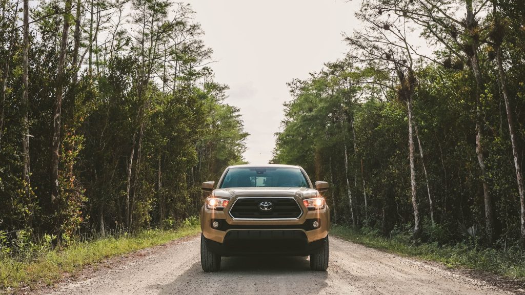 A Beige Toyota driving down the middle of a dirt road with tall trees on each side.